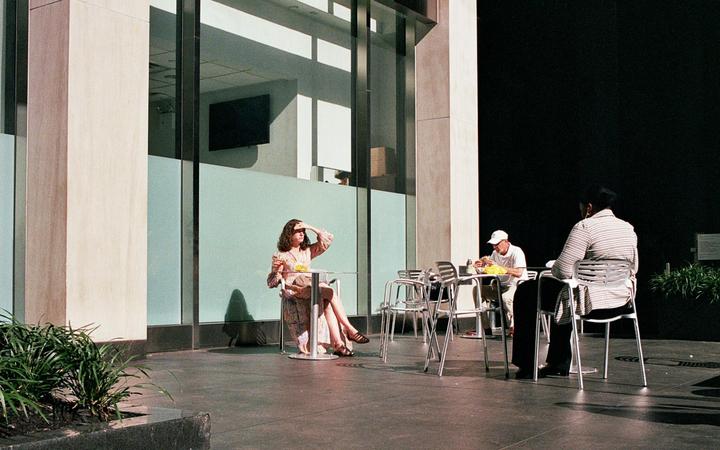 A woman shields her eyes from the sun while sitting at an outdoor table with two men.