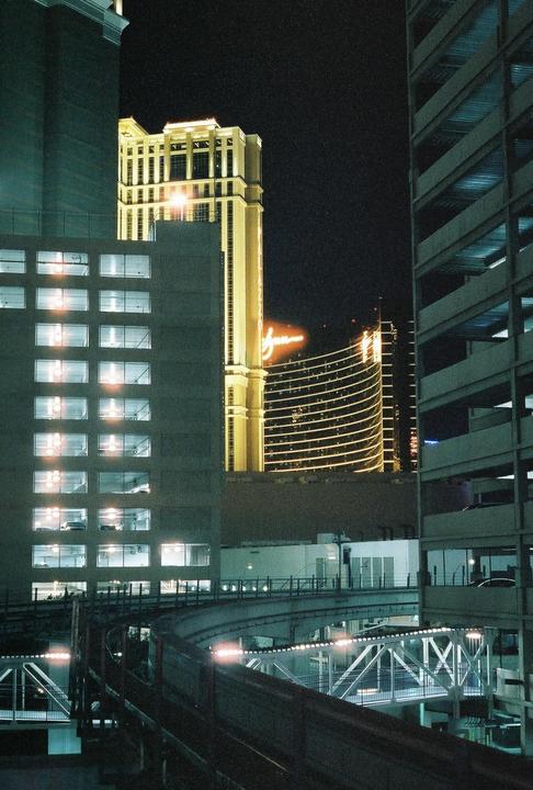 A monorail track curves through a city at night, with brightly lit buildings and a parking garage visible.