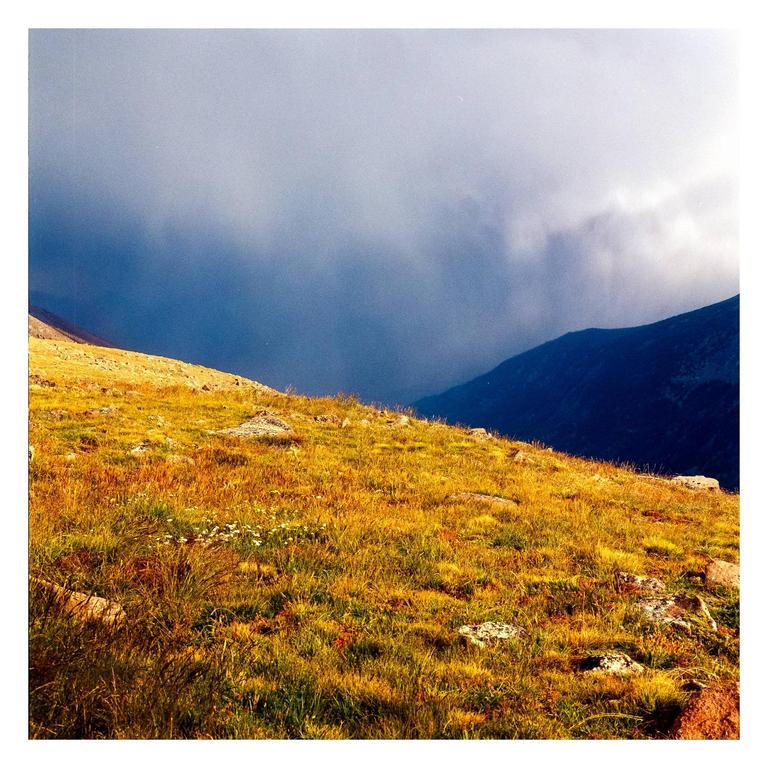 A mountain landscape shows a grassy hillside in the foreground and a dark, stormy sky over distant mountains.