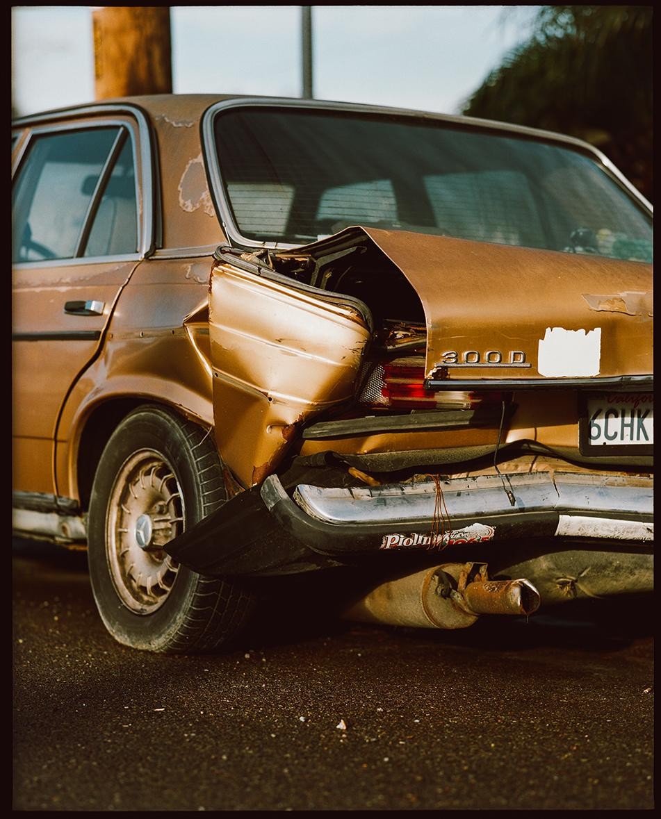 The rear of a gold sedan with significant damage to its trunk and bumper is parked on an asphalt surface.