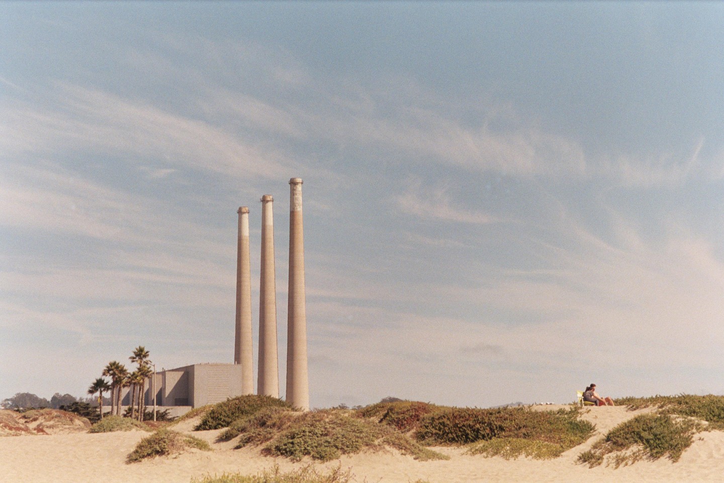 Three tall smokestacks rise behind a sandy beach with a person sitting on a dune.