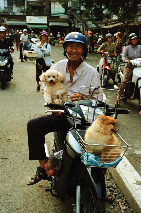 A man on a scooter smiles at the camera while holding a small dog and another dog sits in a basket on the front of the scooter.