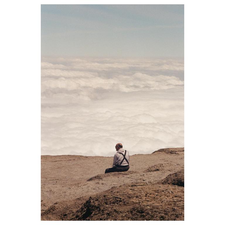 A man sits on a rocky cliff overlooking a vast expanse of clouds.