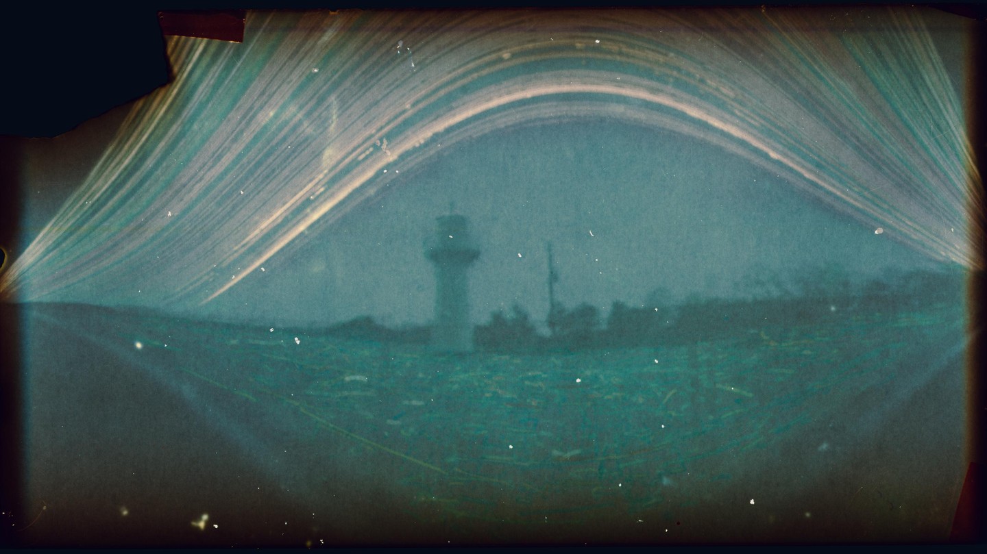 A long exposure image shows a lighthouse on a distant shore under an arched sky with light trails.