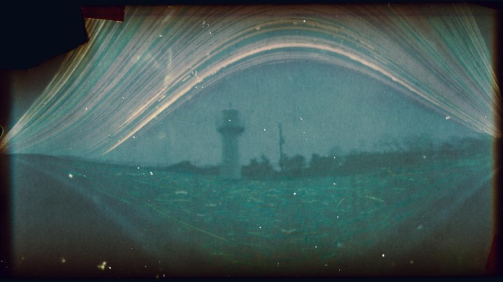 A long exposure image shows a lighthouse on a distant shore under an arched sky with light trails.