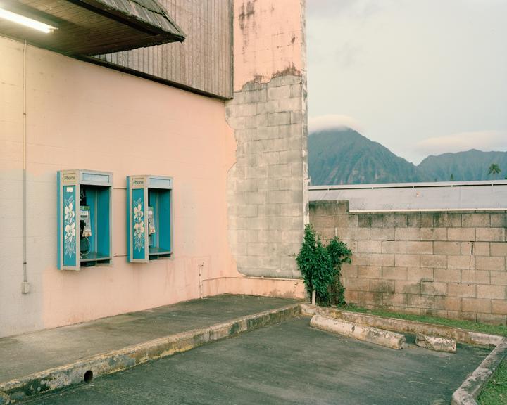 Two payphones are mounted on a pink wall next to a parking lot with a mountain in the background.