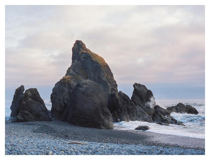Large rock formations stand on a pebble beach with waves crashing against them under a cloudy sky.