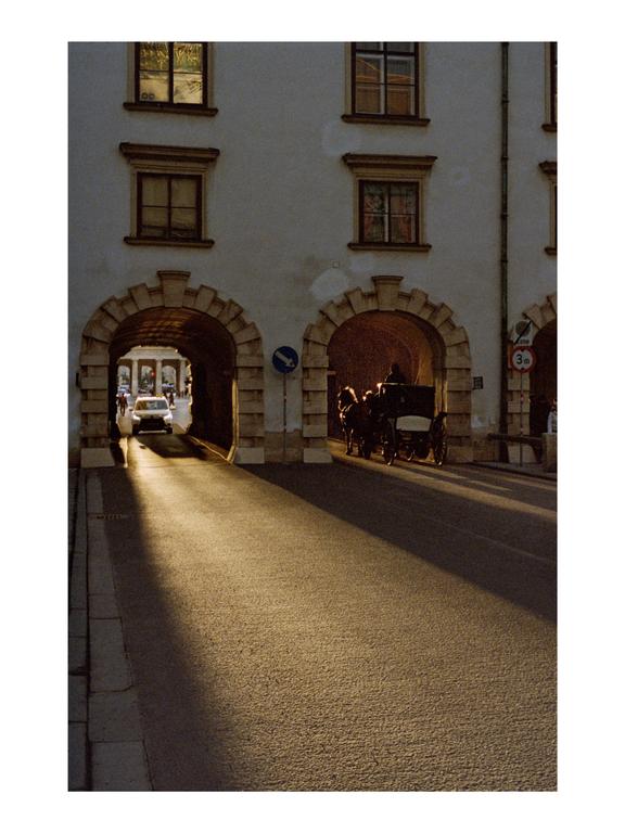 A horse-drawn carriage and a car are visible through two archways in a building facade, with a long shadow cast across the street.