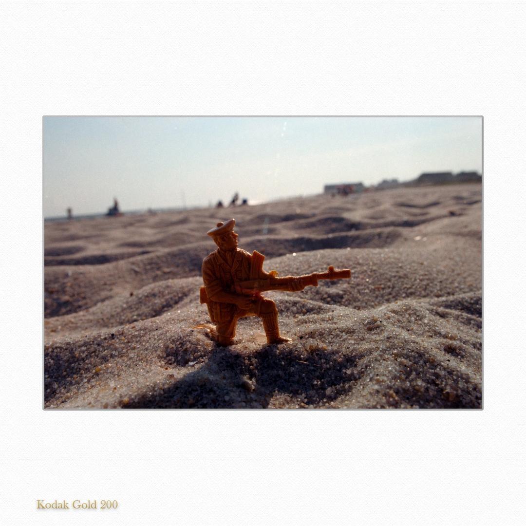 A toy soldier kneels in the sand on a beach with people in the background.
