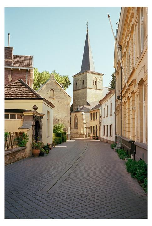 A narrow cobblestone street curves through a historic town with a tall church steeple visible in the background.