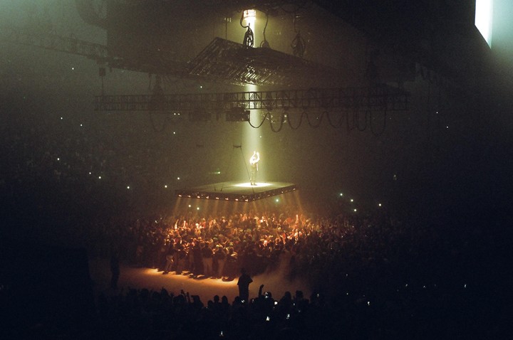 A performer stands on a raised stage under a spotlight, surrounded by a large crowd in a dark arena.