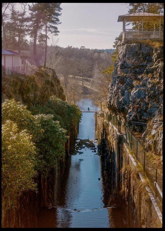 A narrow river flows between two steep, rocky banks, with a small bridge and a person visible in the distance.