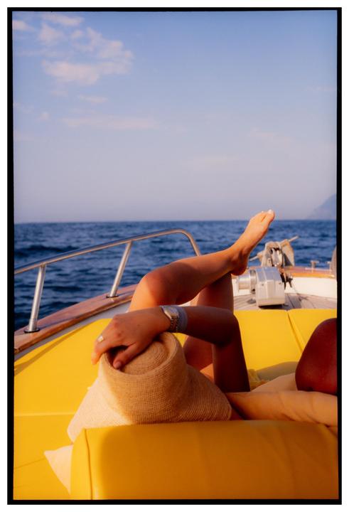A woman relaxes on a boat with her legs crossed and a straw hat on her lap.