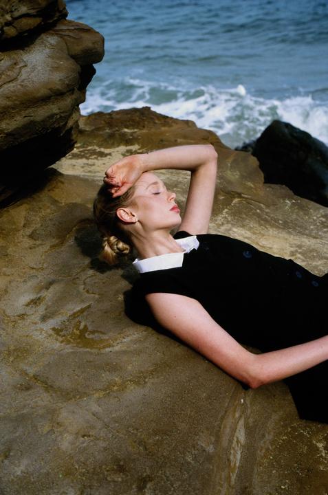 A woman in a black dress with a white collar lies on a rock by the ocean.