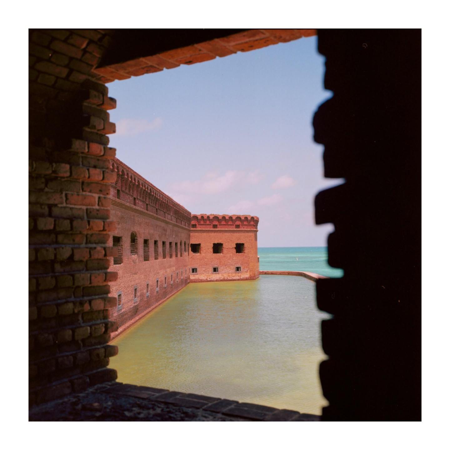 A brick fort with a moat is viewed through a dark, rectangular opening, with the ocean visible in the distance.
