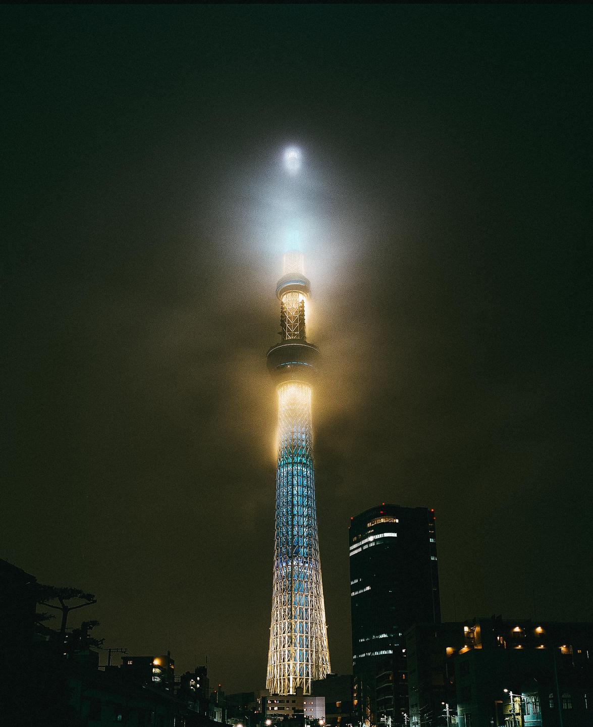 A tall, illuminated tower stands shrouded in fog at night, with other buildings visible below.