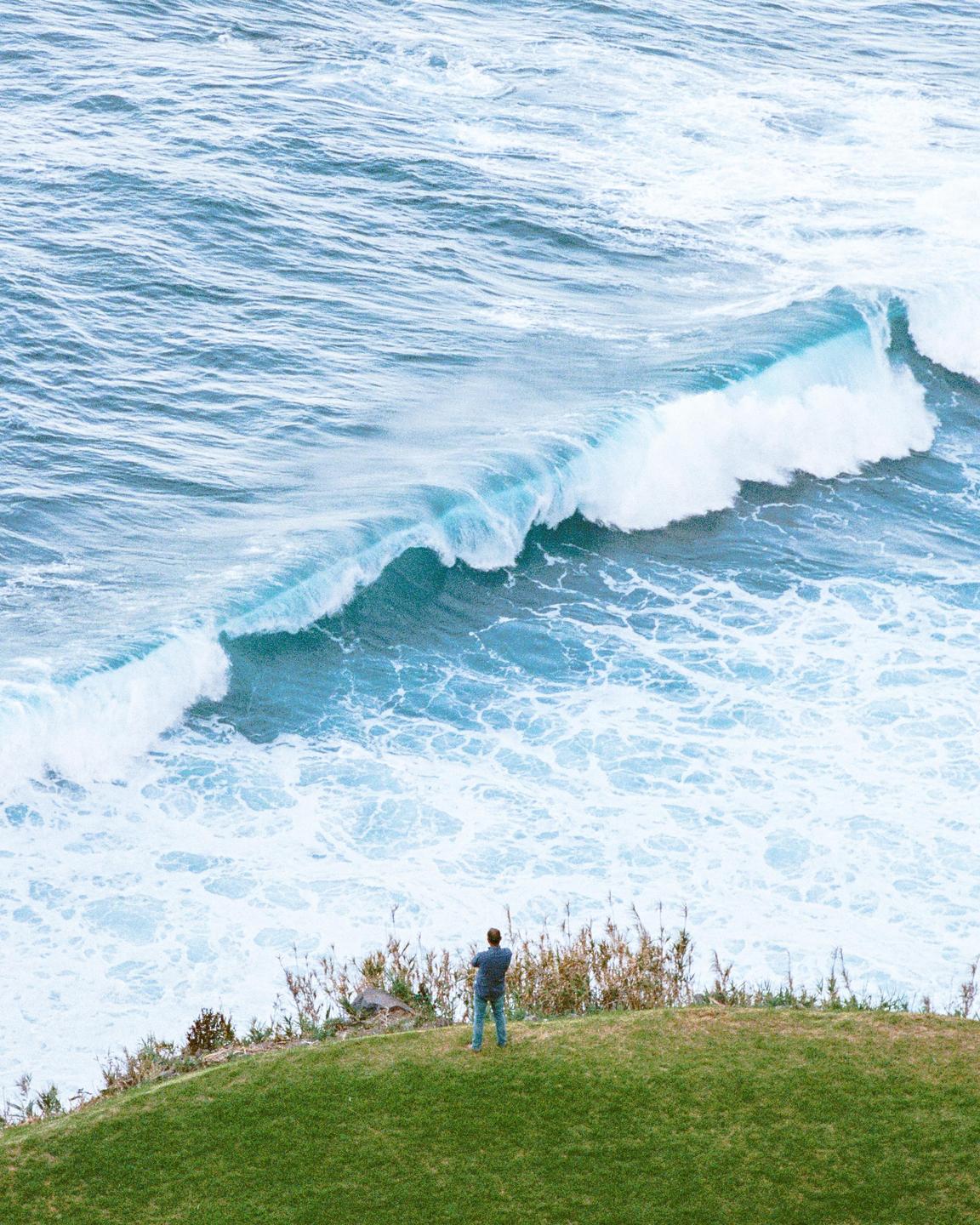 A man stands on a grassy cliff overlooking a large ocean wave.
