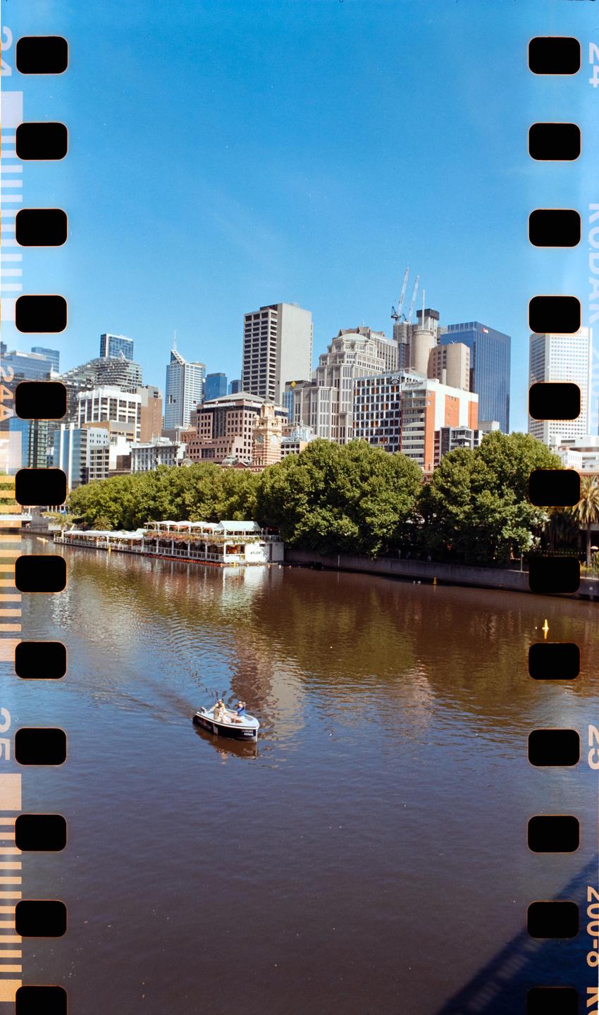 A small boat with two people floats on a river with a city skyline and trees in the background.