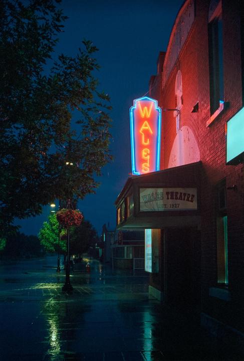A neon sign for the Wales Theatre glows red and blue on a brick building next to a tree on a wet street at night.