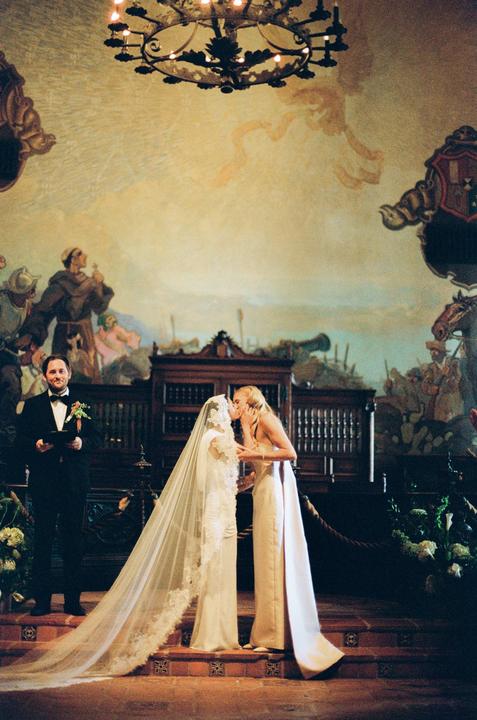 A woman in a wedding dress and veil kisses another woman in a long dress while a man in a tuxedo stands to the left, all in a room with a painted mural and a chandelier.