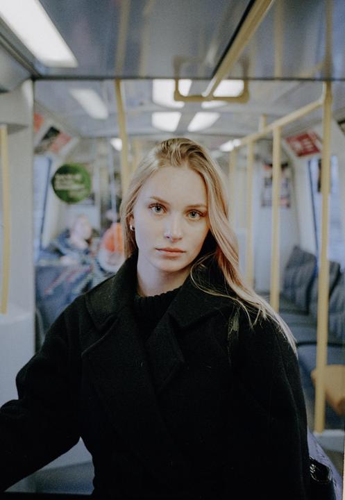 A woman with long blonde hair wears a black coat and sweater while riding a subway.