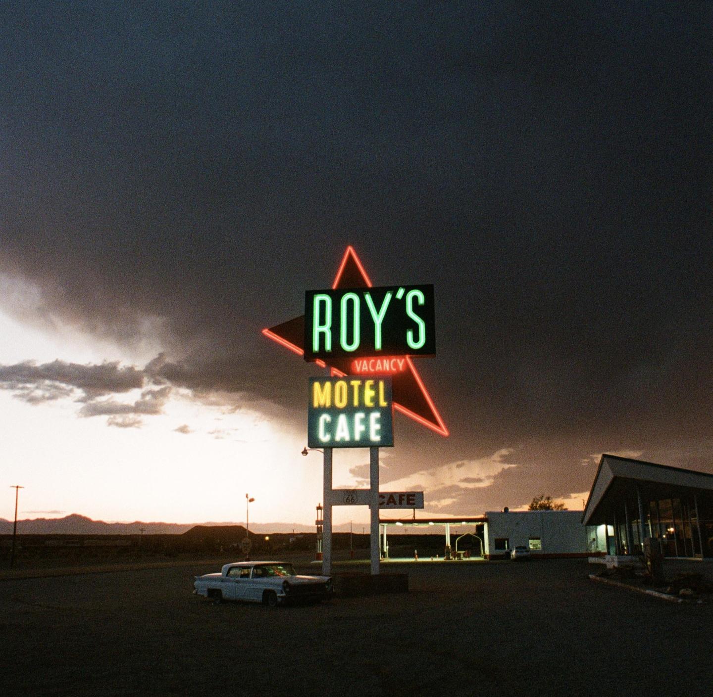 A neon sign for Roy's Motel and Cafe stands illuminated against a dark, cloudy sky at dusk, with a vintage car parked in the foreground.