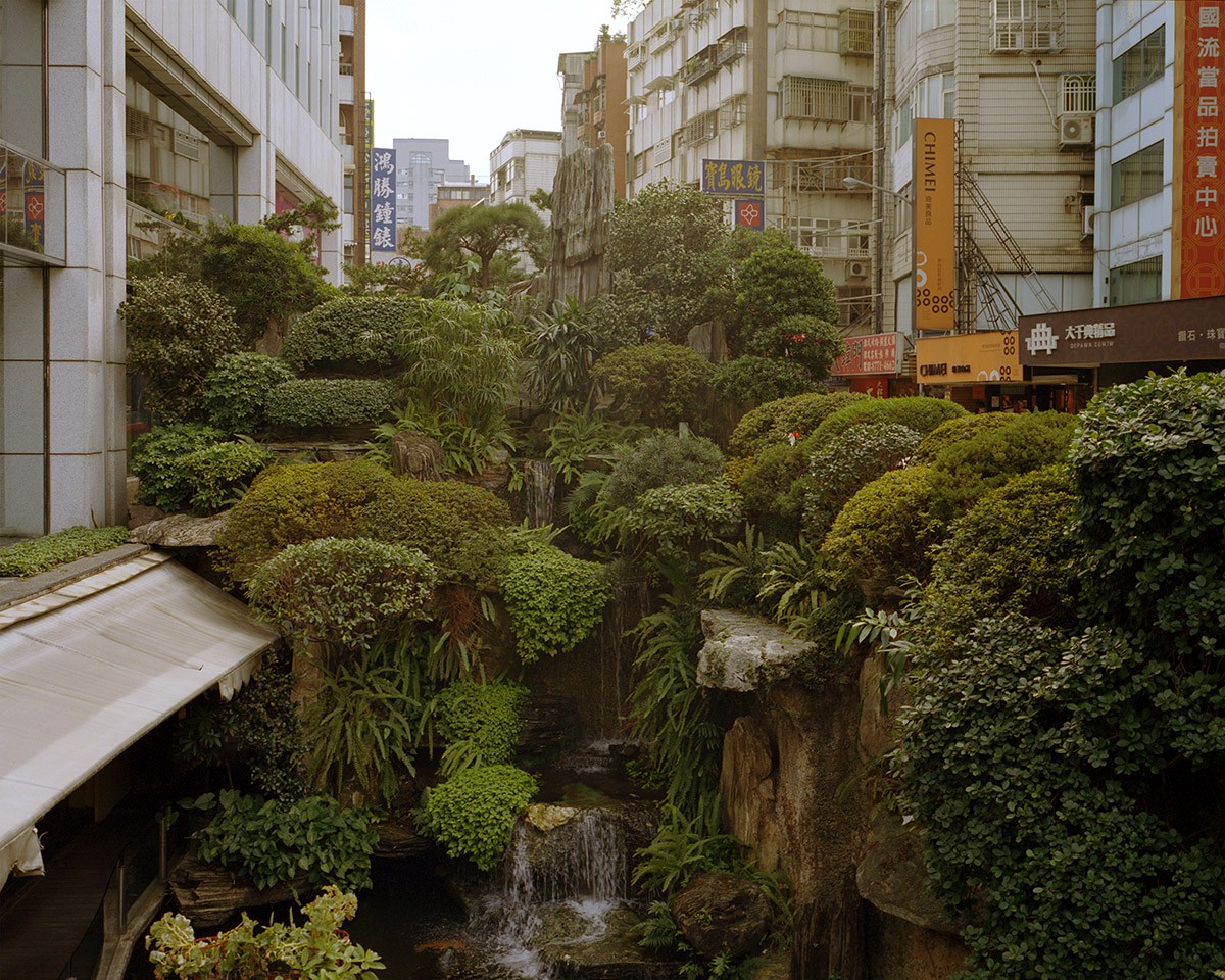 A lush, multi-tiered garden with waterfalls is nestled between tall city buildings with various signs.