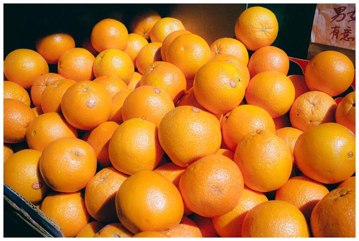A large pile of oranges is displayed in a box, with some Chinese characters visible on a sign in the upper right corner.
