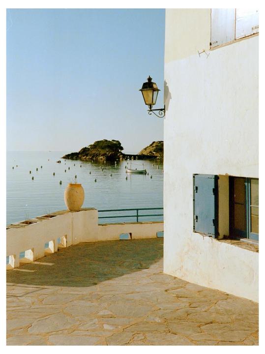 A stone patio with a white wall and blue-shuttered window overlooks a calm bay with small islands and a boat.