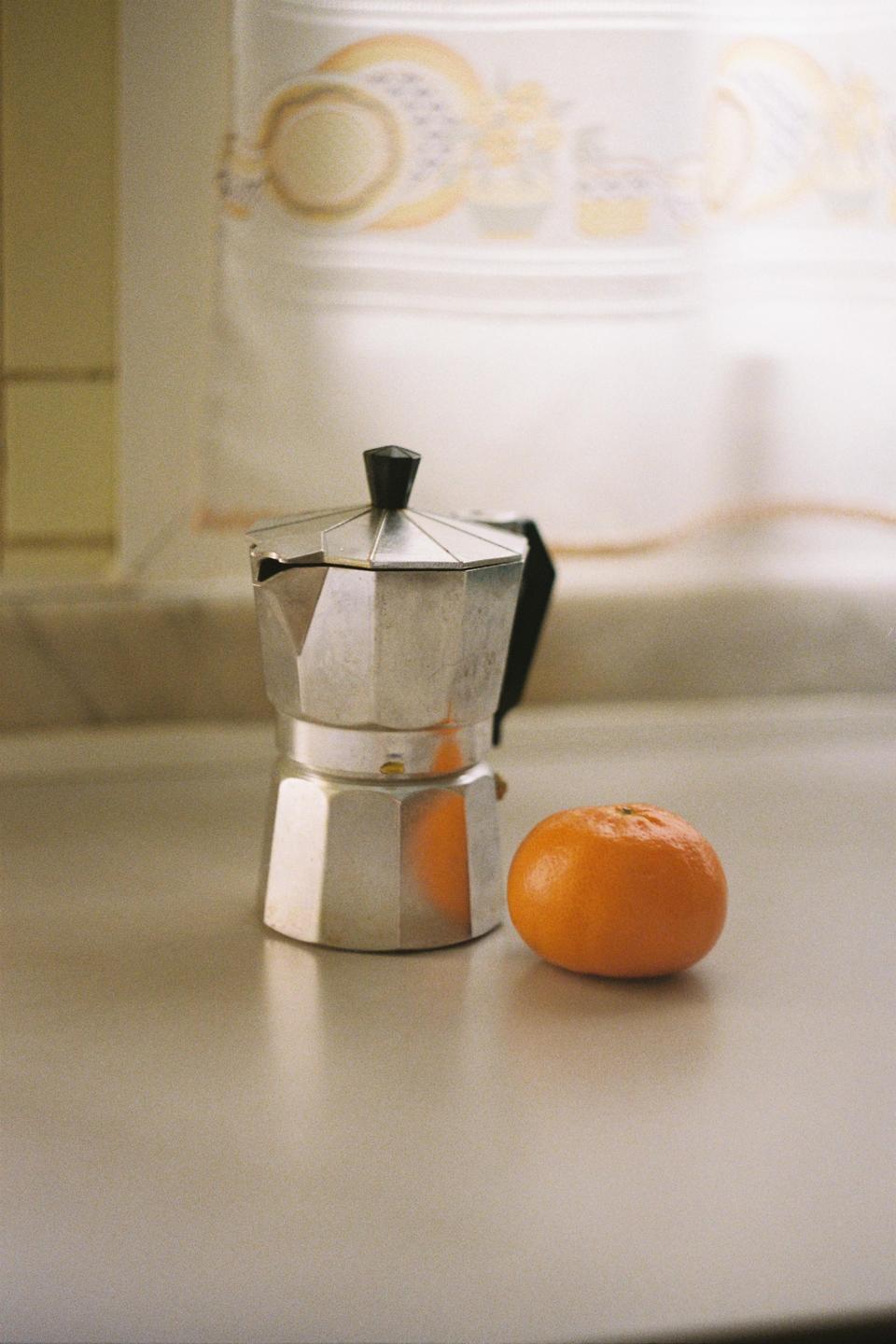 A moka pot and a tangerine sit on a counter in front of a patterned curtain.