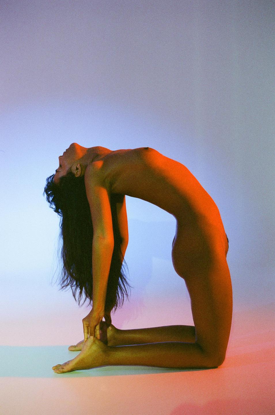 A woman with long hair is kneeling and bending backward in a yoga pose with colorful lighting.