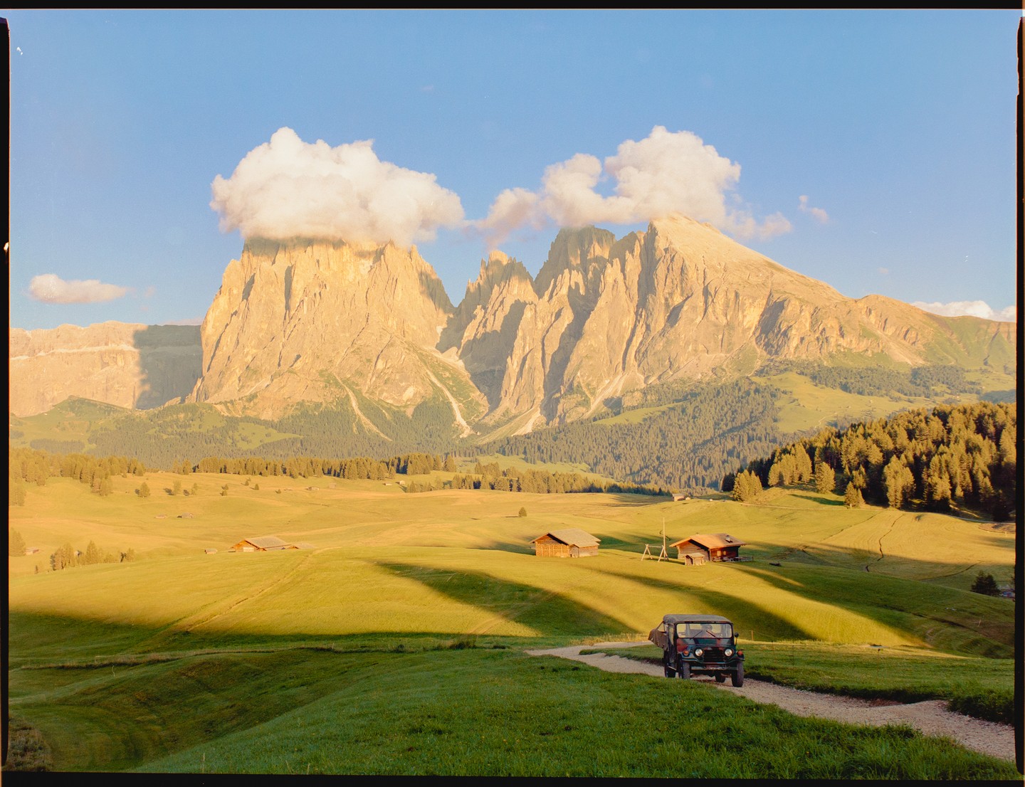 A jeep drives on a dirt road through a green valley with mountains and clouds in the background.