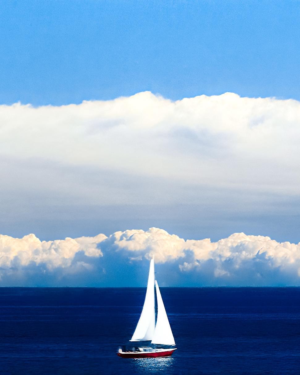 A red sailboat with white sails floats on a deep blue sea under a bright blue sky with a band of white clouds.