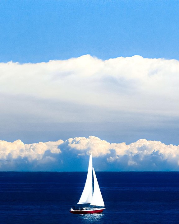 A red sailboat with white sails floats on a deep blue sea under a bright blue sky with a band of white clouds.