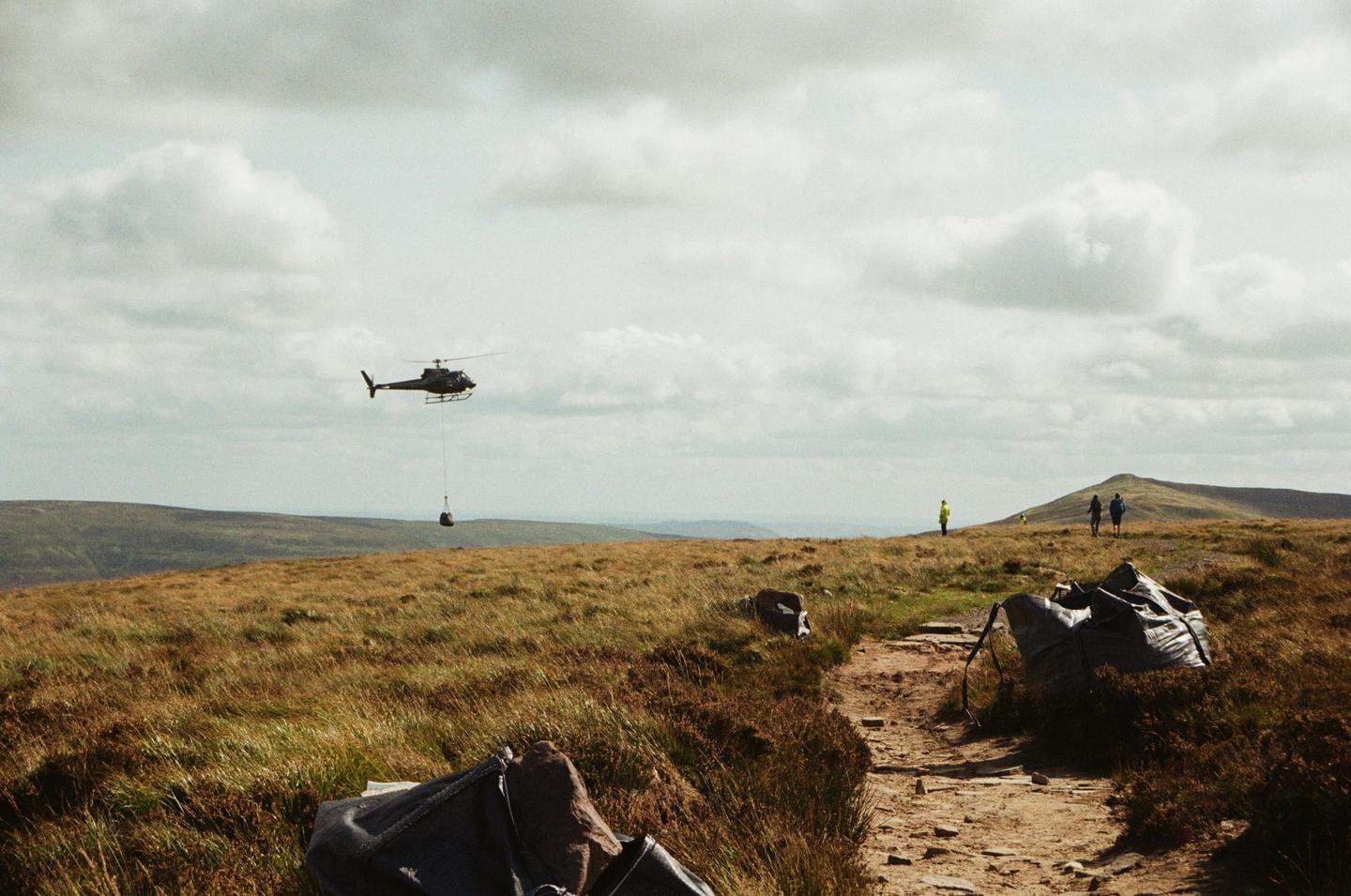 A helicopter carries a load over a grassy hill where people are walking on a dirt path.