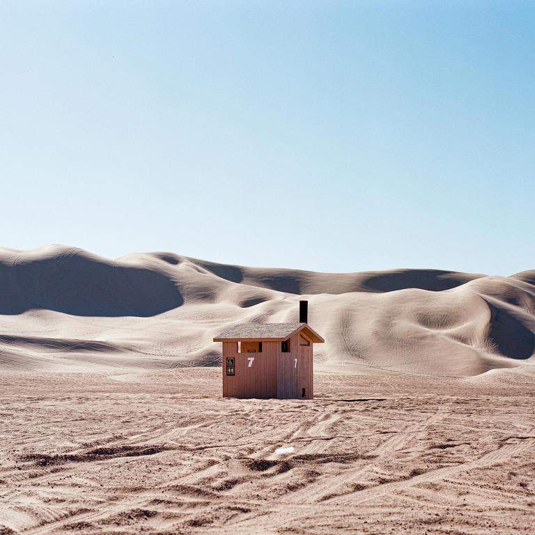 A small wooden building with a dark chimney stands in a vast desert landscape with sand dunes in the background.