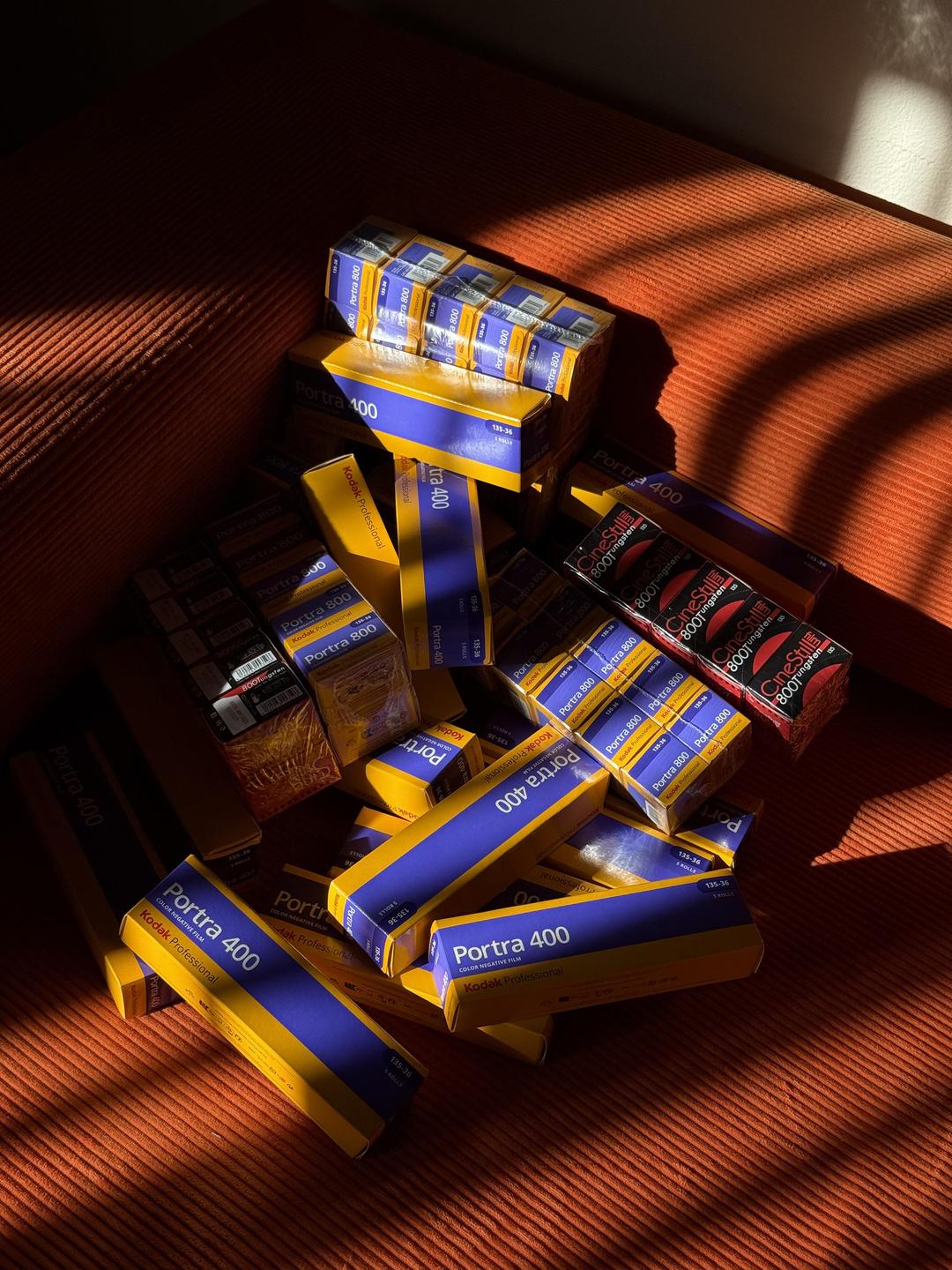 A pile of film boxes sits on an orange textured surface, illuminated by sunlight creating strong shadows.