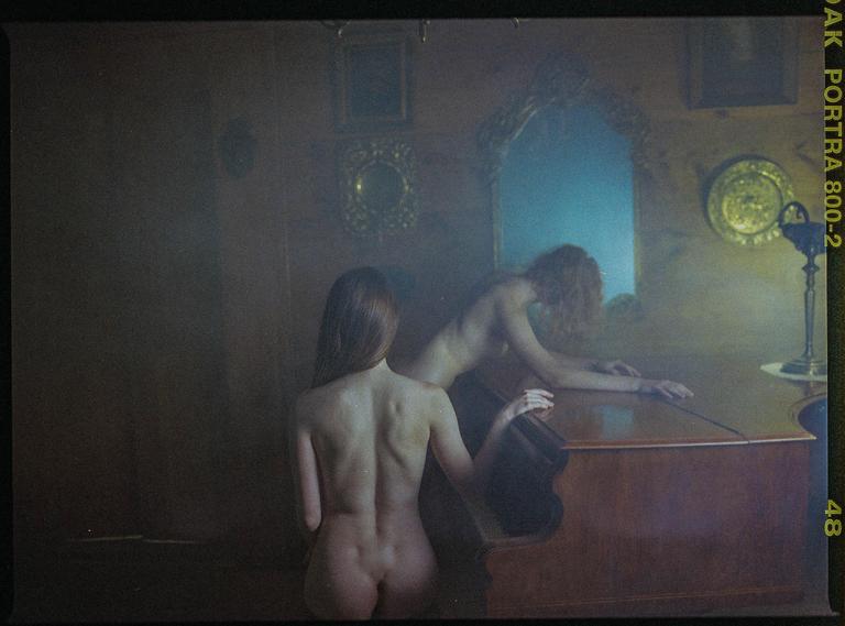 Two nude women are in a dimly lit room with one woman sitting in front of a piano and the other leaning over it.