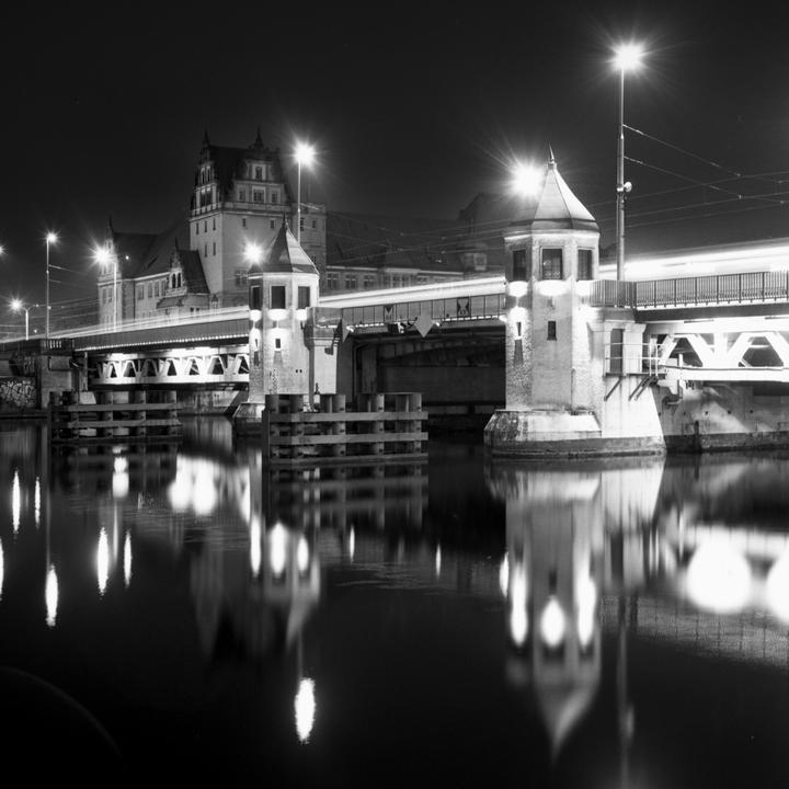 A bridge with two towers and a building in the background is illuminated at night, with reflections visible in the water below.