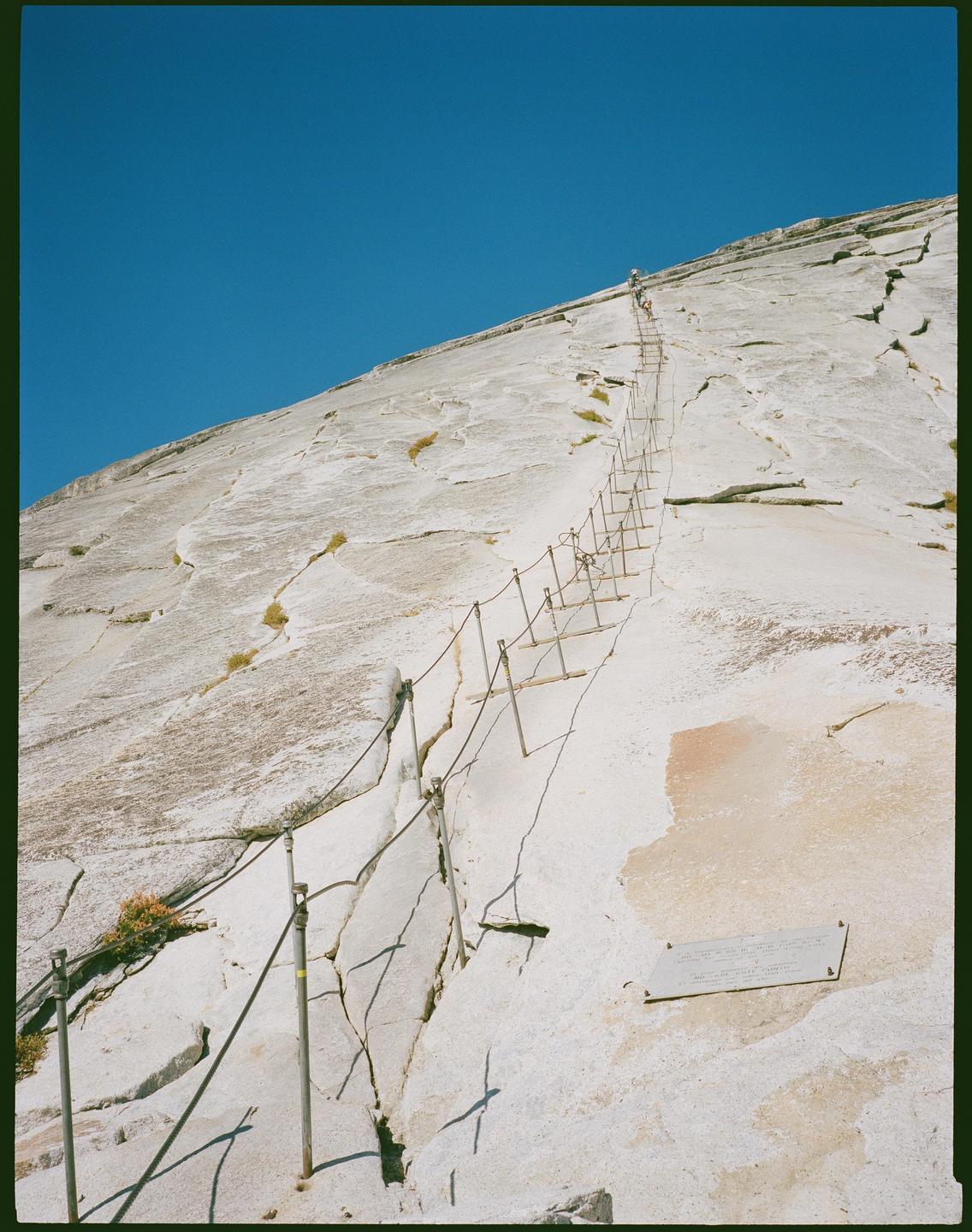 A steep cable route ascends a large granite dome under a clear blue sky.