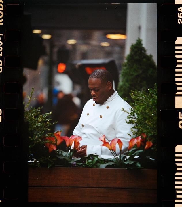 A man in a white chef's coat looks down at his phone while standing behind a planter with orange flowers.