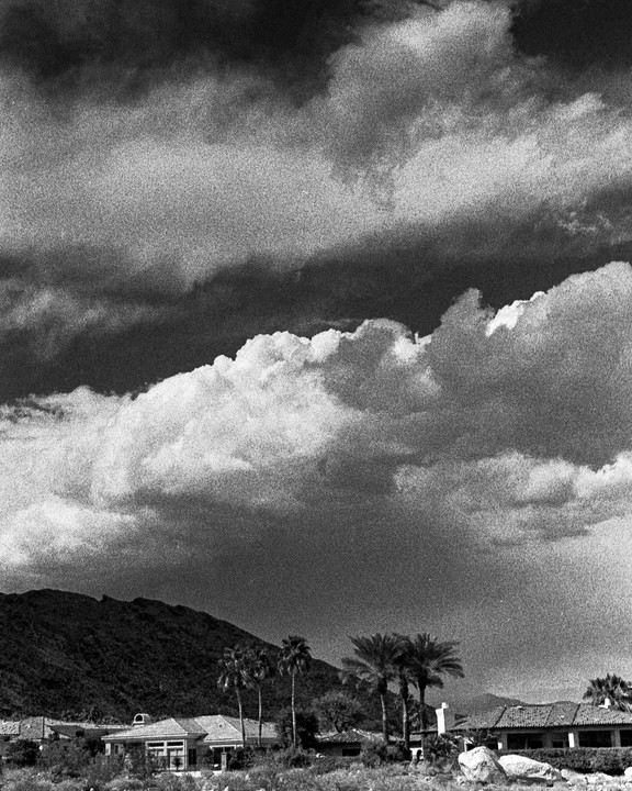Dark clouds hang over a desert landscape with palm trees, houses, and a mountain.