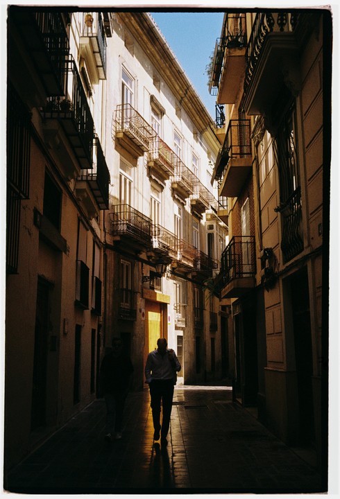 Two men walk down a narrow street lined with tall buildings and balconies, with one man illuminated by a shaft of sunlight.