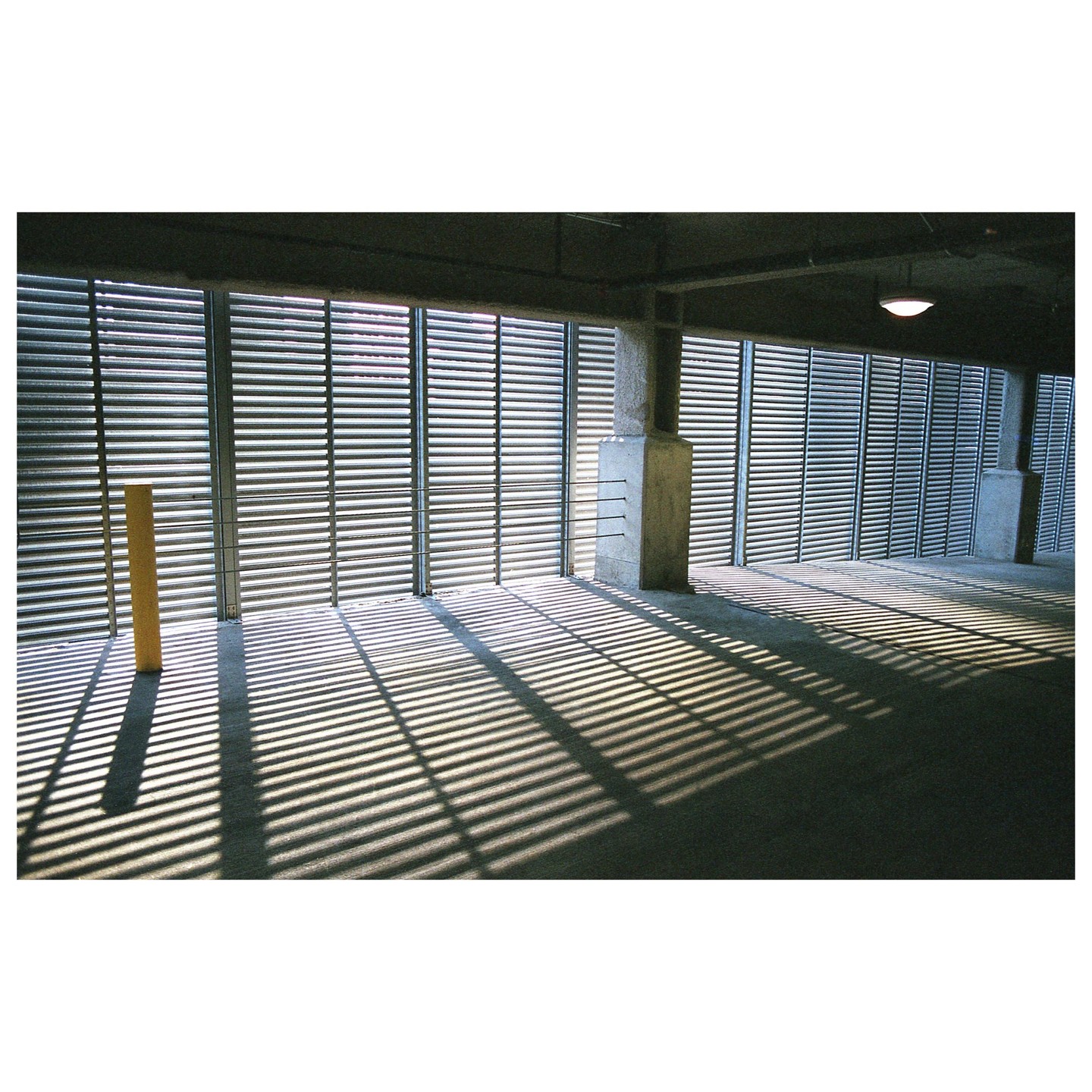 Sunlight streams through horizontal blinds, casting long, striped shadows across a concrete floor in a parking garage.