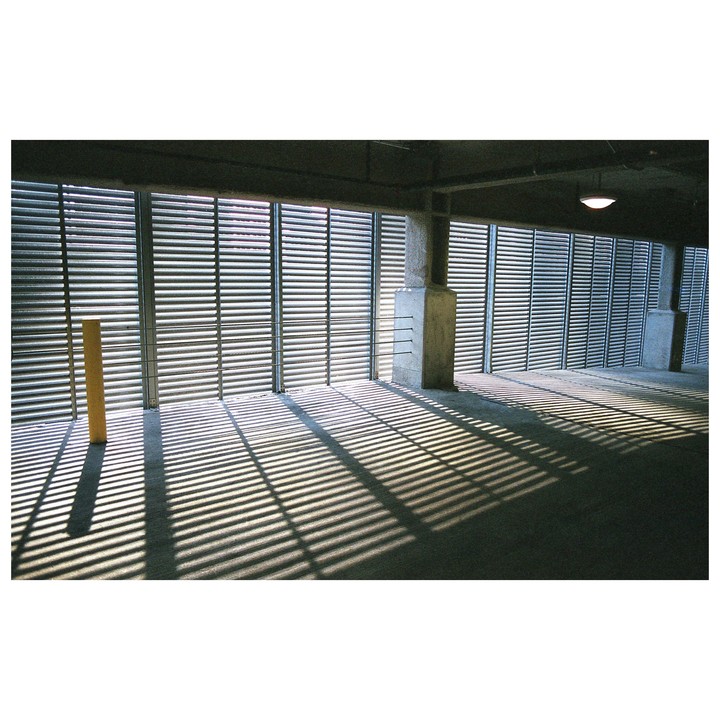 Sunlight streams through horizontal blinds, casting long, striped shadows across a concrete floor in a parking garage.