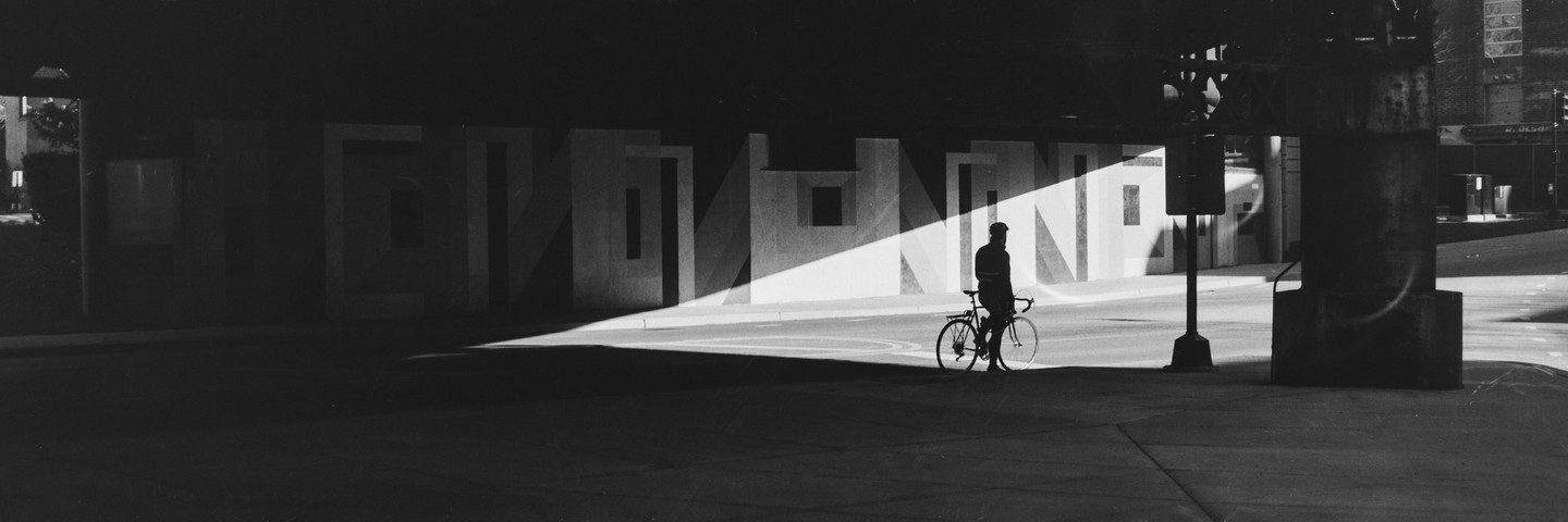 A man stands with his bicycle in a shaft of light under an overpass.