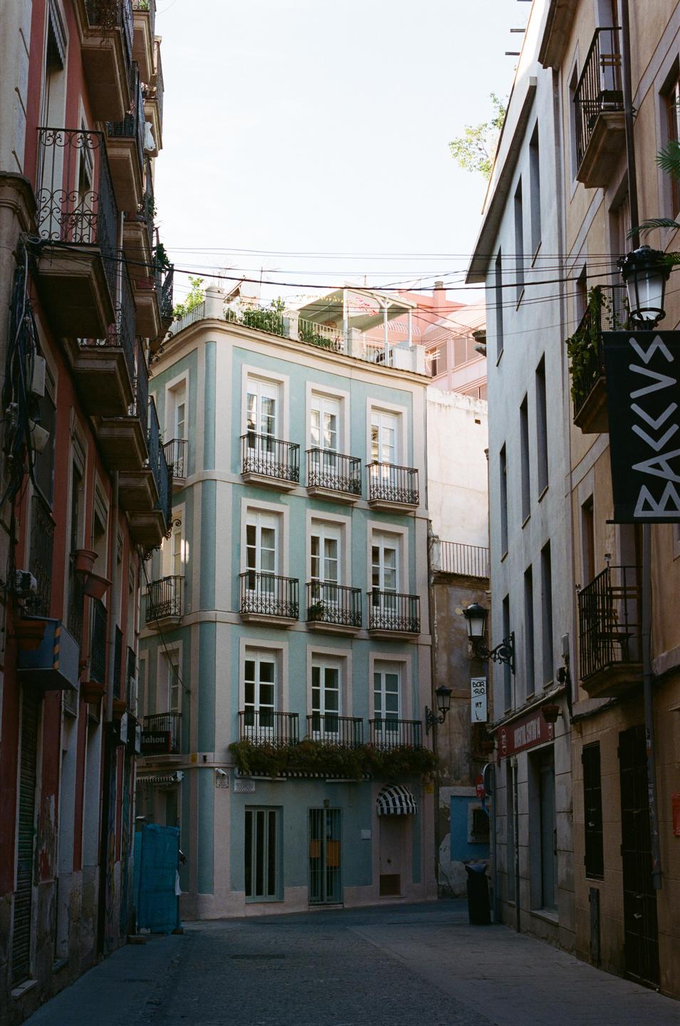 A narrow street is lined with tall buildings featuring balconies and a light blue corner building.