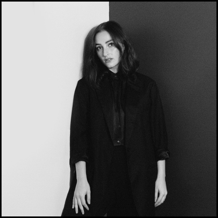 A woman in a dark shirt and jacket stands against a wall that is half white and half black.