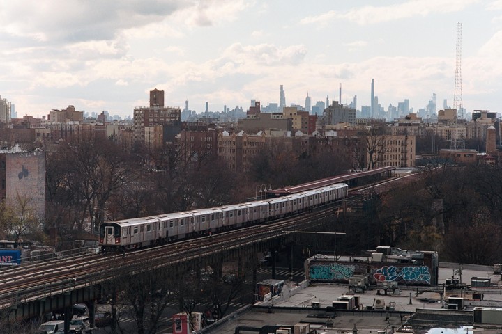 A subway train travels on an elevated track through a city with a skyline in the distance.