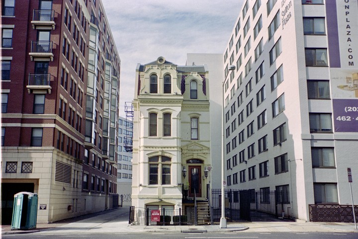 A narrow, ornate white building stands between two larger, modern buildings on a city street.